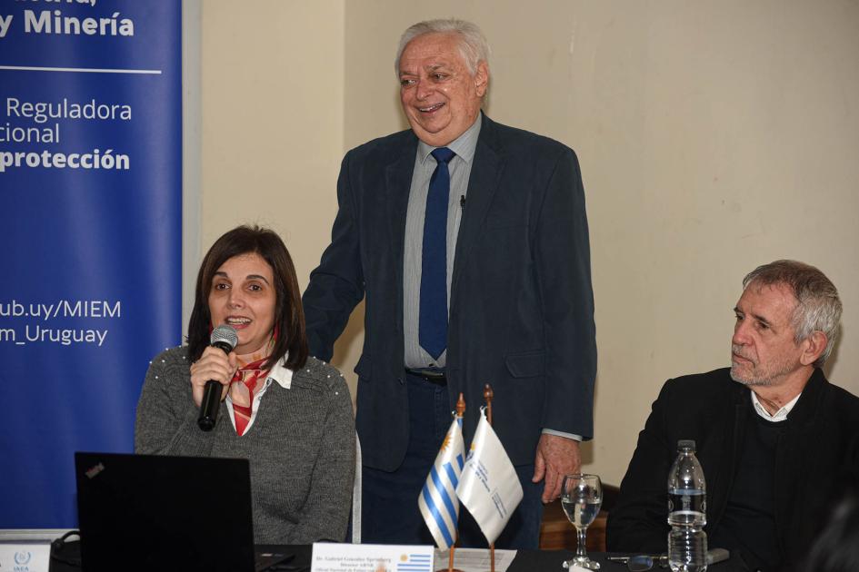La Dra. Virginia Roldós, experta local de la ARNR, en la jornada de la apertura Una mujer habla con un micrófono, en una mesa; a su lado, un hombre sentado y otro de pie