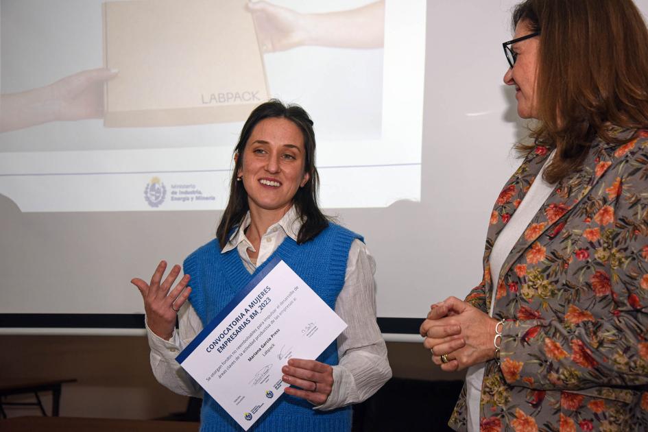 Mariana García Press explicó cómo aumenta su productividad con el software adquirido con los fondos Mariana García Press sostiene su diploma mientras dialoga con Facio; detrás, pantalla