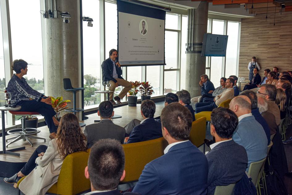 Juan Urthiague, de Globant Público, de espaldas, escucha a un hombre en un escenario; a su lado, la moderadora
