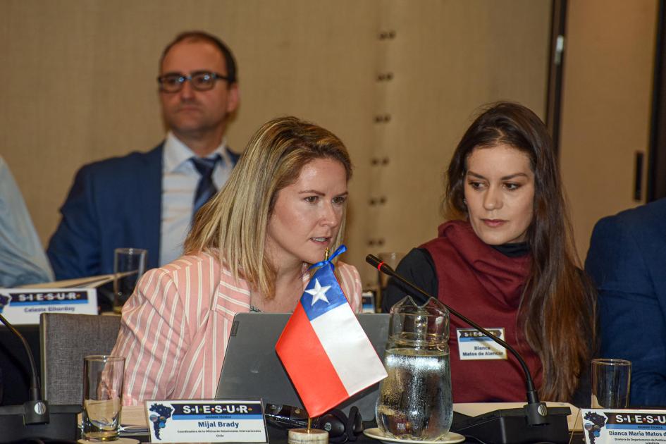 Representantes de los países integrantes de SIESUR Mujer habla al lado de otra; en su mesa hay una bandera de Chile