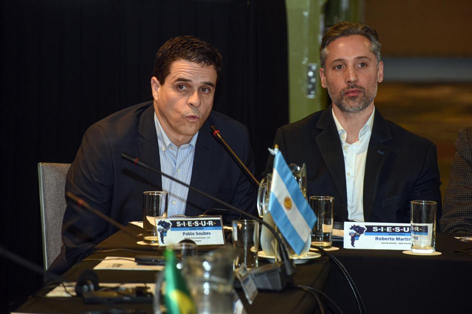 Representantes de los países integrantes de SIESUR Hombre habla junto a otro; en la mesa, bandera de Argentina