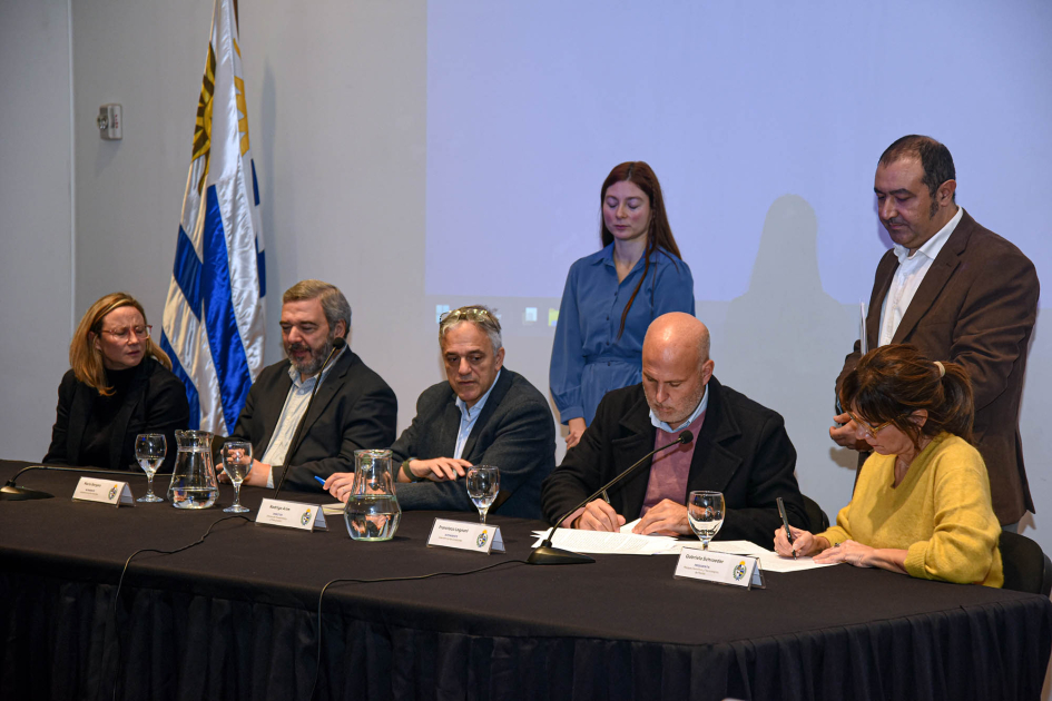 Cinco personas firman el convenio en una mesa; detrás, otras personas, de pie, y bandera uruguaya