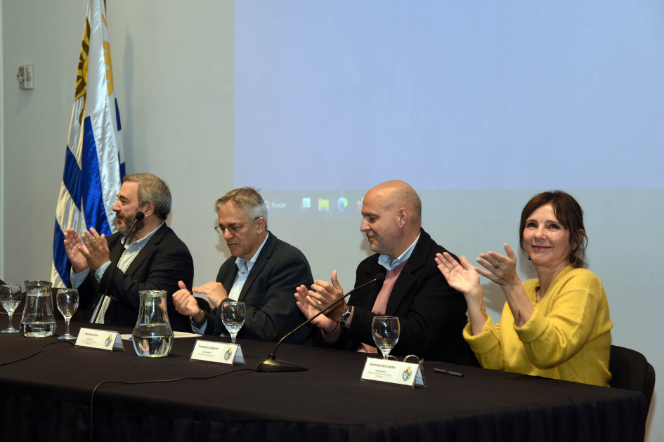 Bergara, Arim, Legnani y Schroeder aplauden; detrás, pantalla y bandera uruguaya