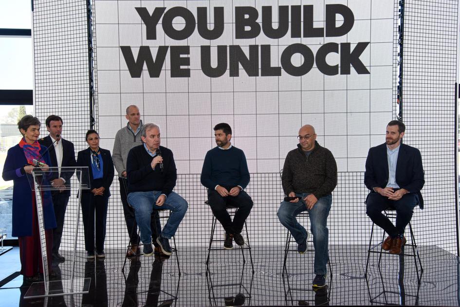Representantes de startups Representantes de las startups hablan desde un escenario; pantalla dice "You build we unlock"