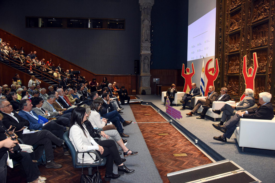 Expositores y expositoras, sentados en sillones; frente, público