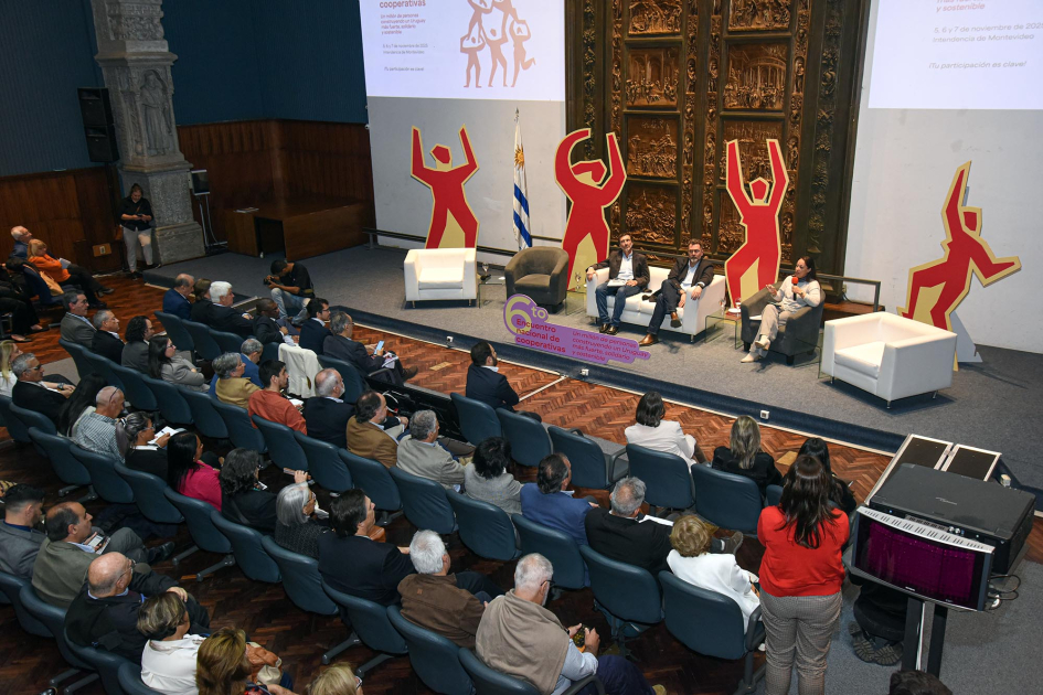 Al fondo, escenario con personas sentadas; más cerca, público, visto de atrás