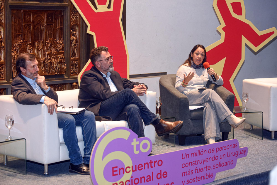 Cardona habla junto a dos hombres; frente a ellos, cartel con nombre del encuentro