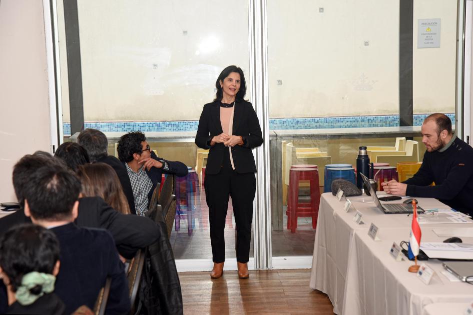 Participantes del taller Participante del encuentro habla, de pie; otros están sentados en mesas paralelas