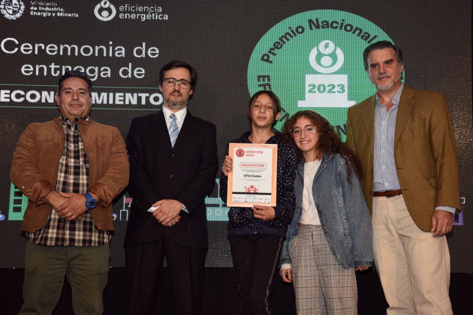 Representantes de la UTU de Colón Tres hombres y dols adolescentes, que sostienen la placa; detrás una pantalla