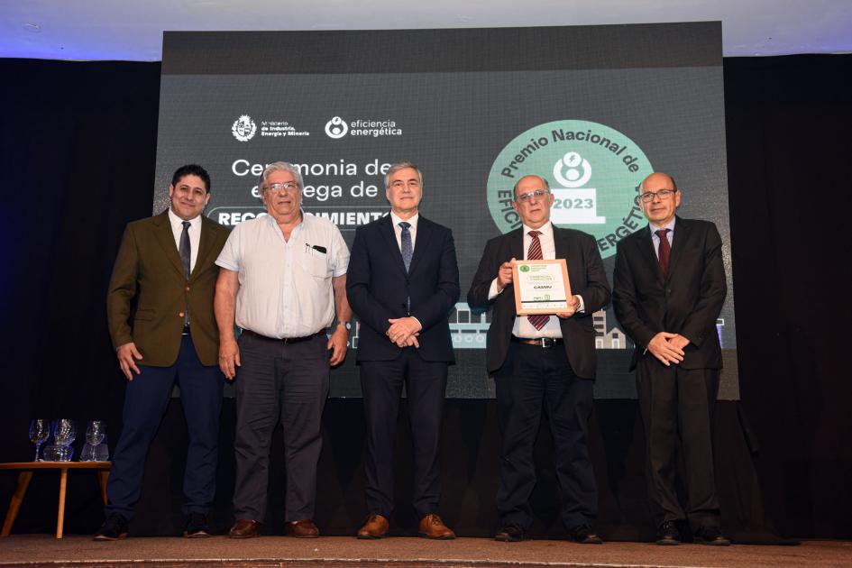 Cinco hombres; uno de ellos sostiene una placa; detrás, una pantallaq