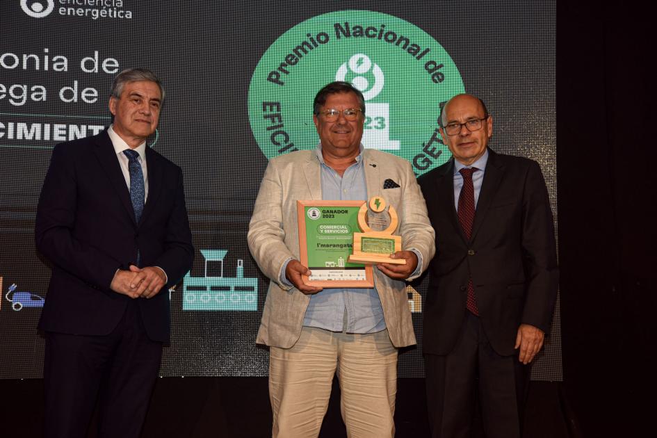 Tres hombres; uno de ellos sostiene el premio y la placa; detrás, una pantalla