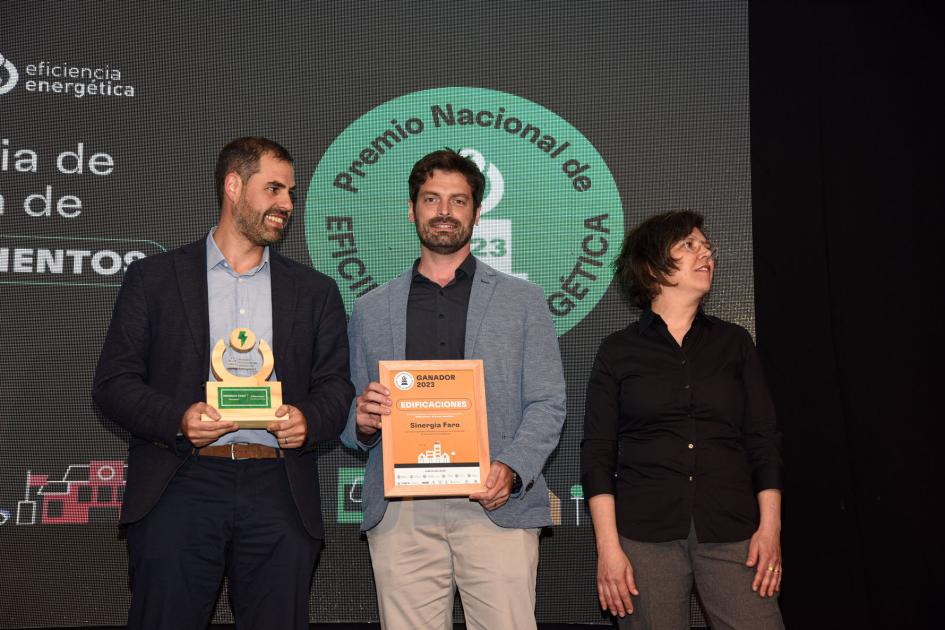 Dos hombres (uno de ellos sostiene el premio y la placa); detrás, una pantalla