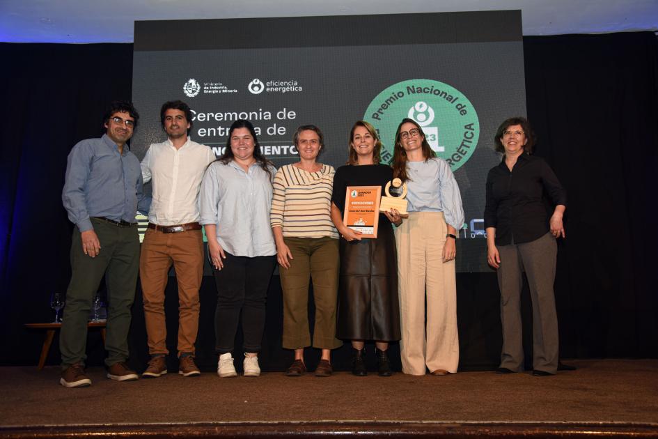 Cinco mujeres (una sostiene el premio y la placa) y dos hombres; detrás hay una pantalla