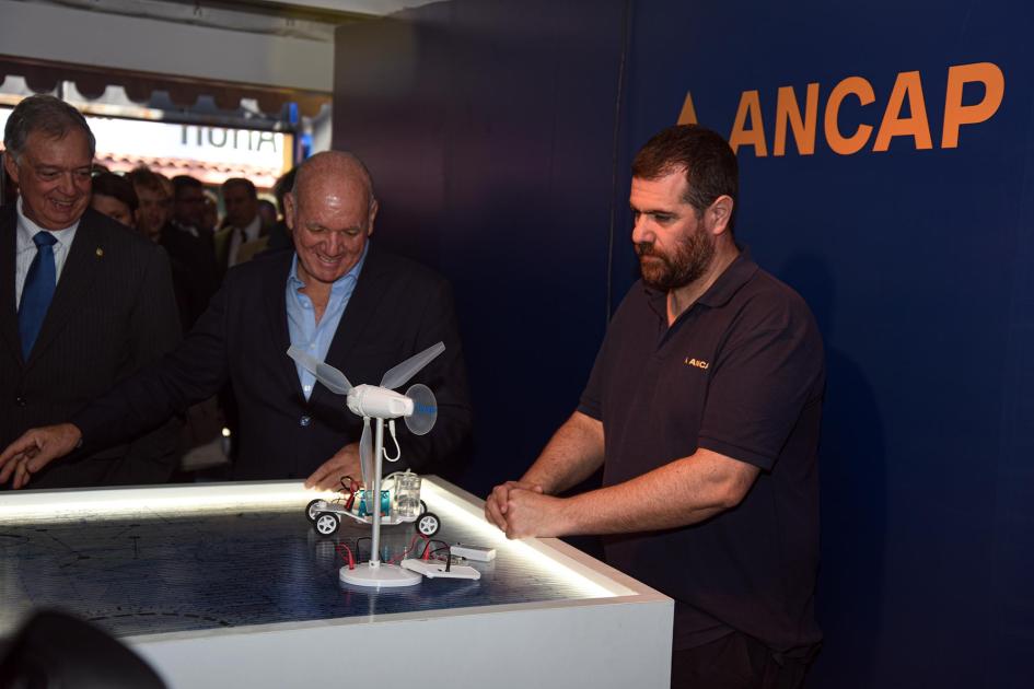 Recorrida del ministro interino Walter Verri en el stand de Ancap Verri observa un robot; junto a él, una persona; detrás, logo de Ancap