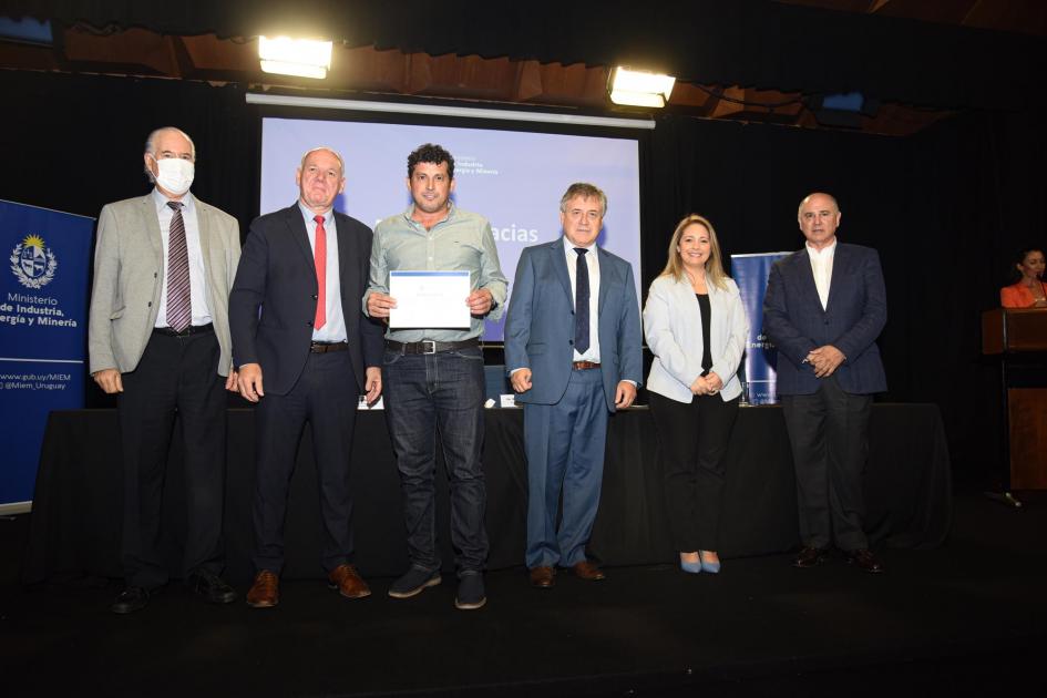 Un hombre muestra su certificado junto a las autoridades; detrás, una pantalla azul