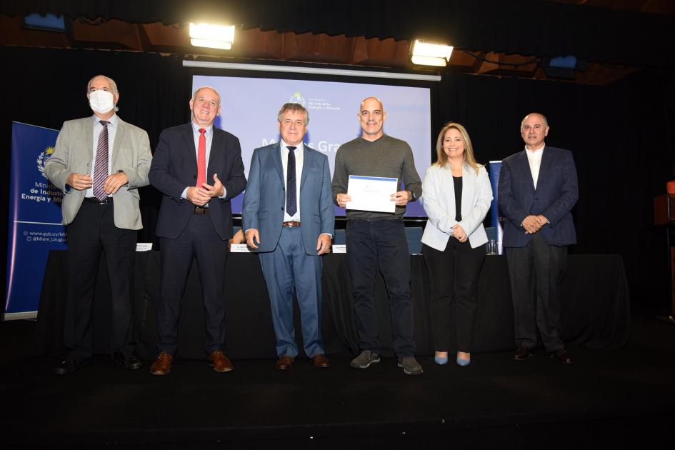 Un hombre muestra su certificado junto a las autoridades; detrás, una pantalla azul