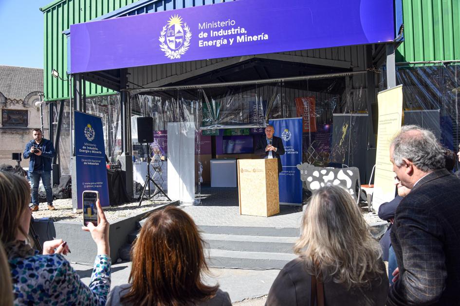Inauguración del stand del MIEM Verri habla desde un atril frente al stand del MIEM, de espaldas, público de pie