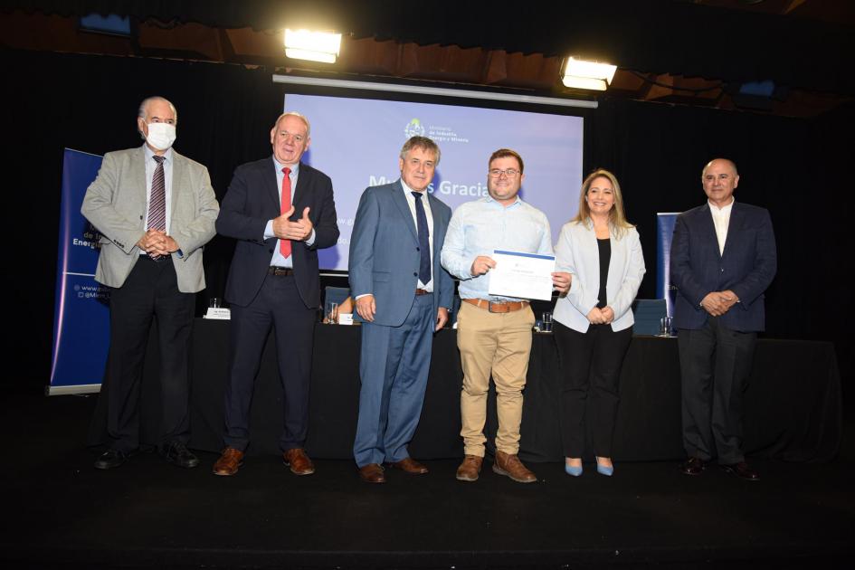 Certificado para Enfoque Publicitario Un hombre muestra su certificado junto a las autoridades; detrás, una pantalla azul