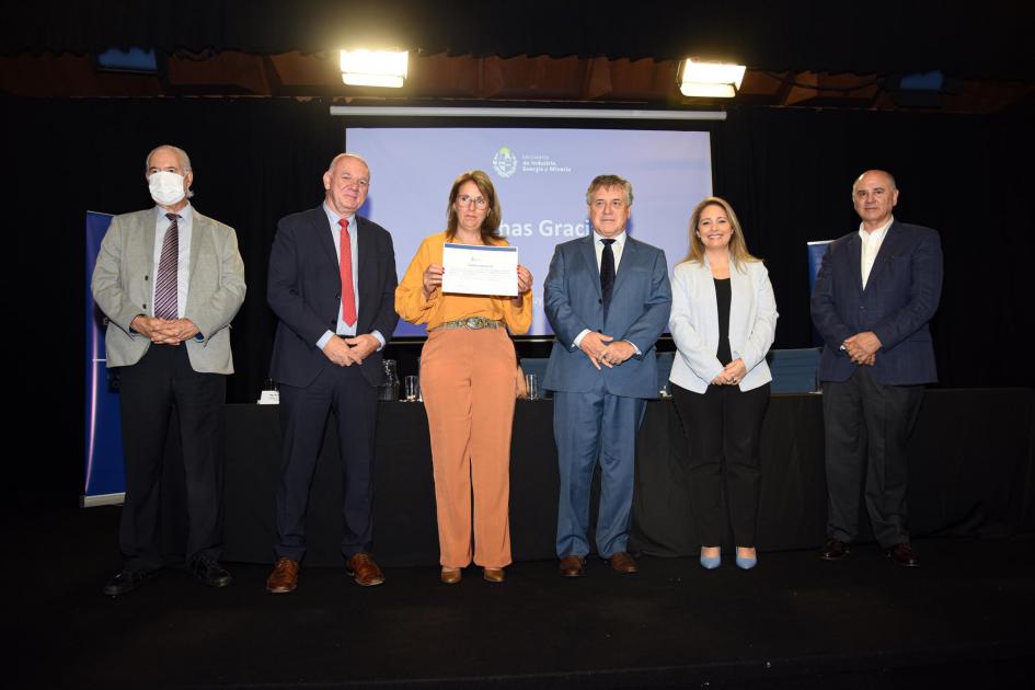 Una mujer muestra su certificado junto a las autoridades; detrás, una pantalla azul