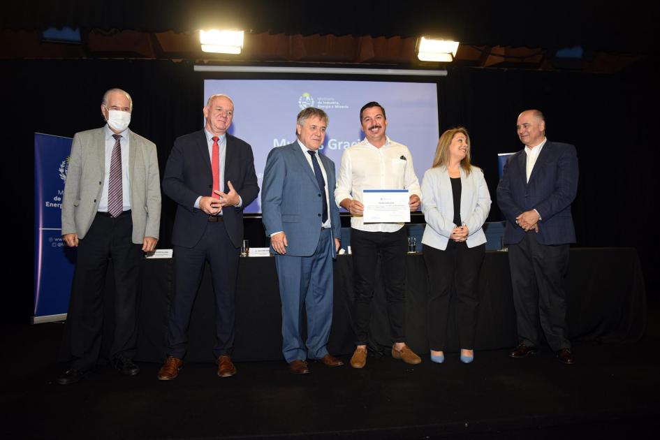 Un hombre muestra su certificado junto a las autoridades; detrás, una pantalla azul