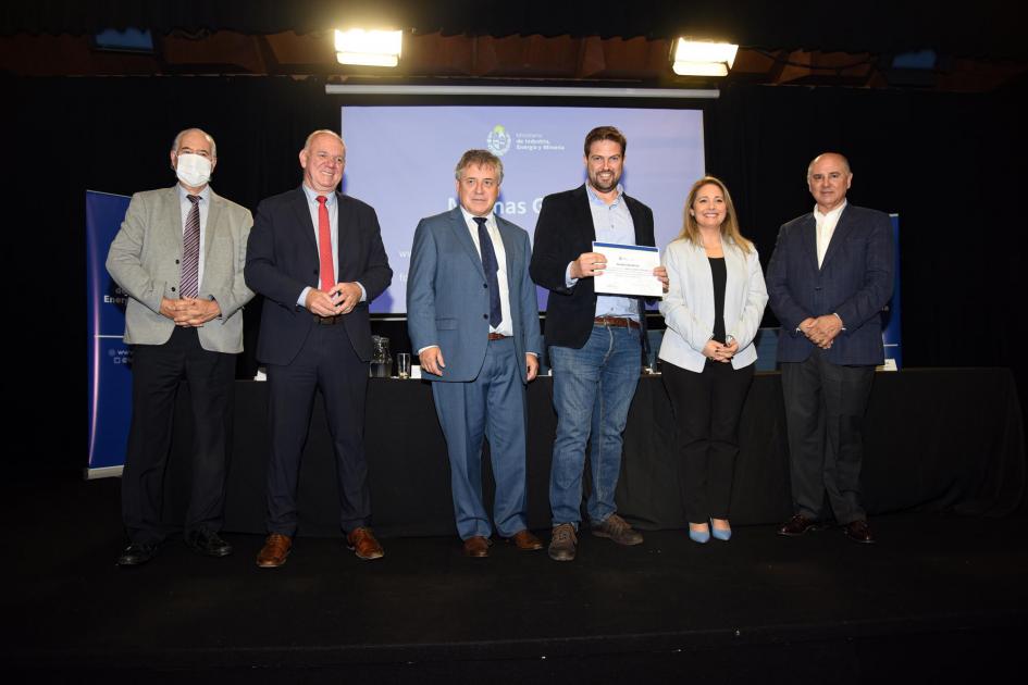 Certificado para Esteban Urban Un hombre muestra su certificado junto a las autoridades; detrás, una pantalla azul