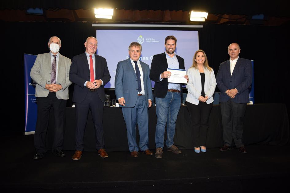 Un hombre muestra su certificado junto a las autoridades; detrás, una pantalla azul