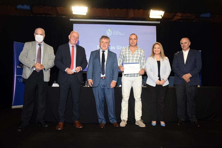 Un hombre muestra su certificado junto a las autoridades; detrás, una pantalla azul