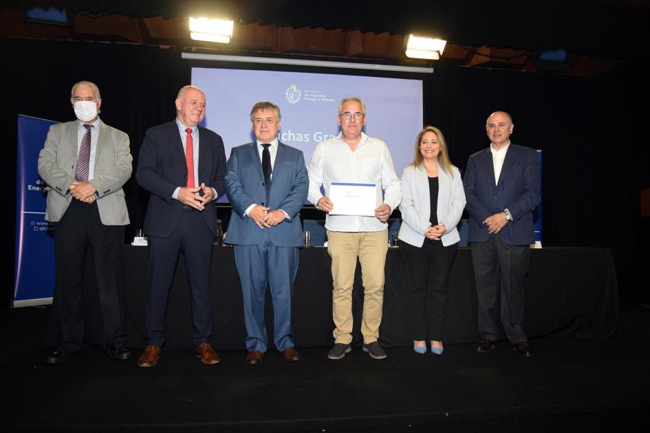 Un hombre muestra su certificado junto a las autoridades; detrás, una pantalla azul