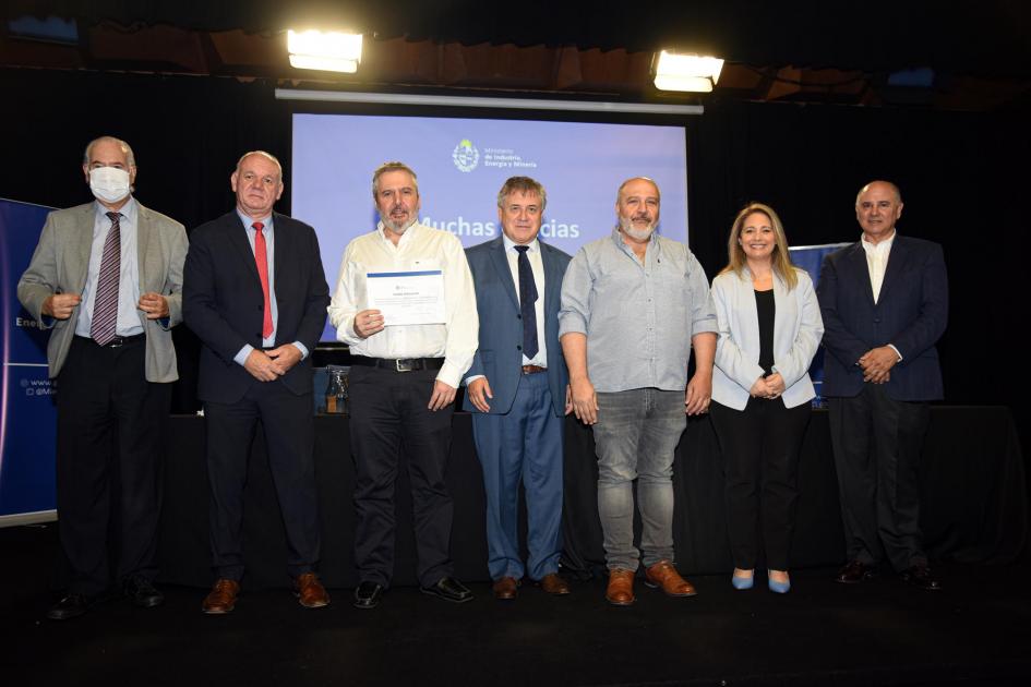 Un hombre muestra su certificado junto a las autoridades y a otro hombre; detrás, una pantalla azul