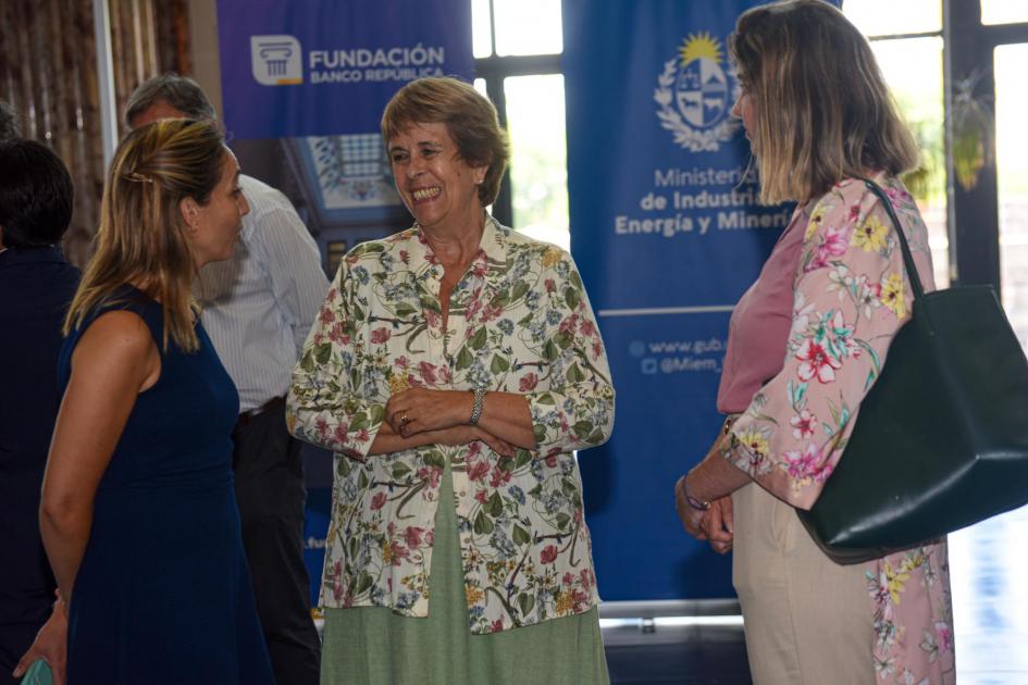 Ana Antía sonríe junto a dos mujeres; detrás, banners azules del MIEM y el BROU