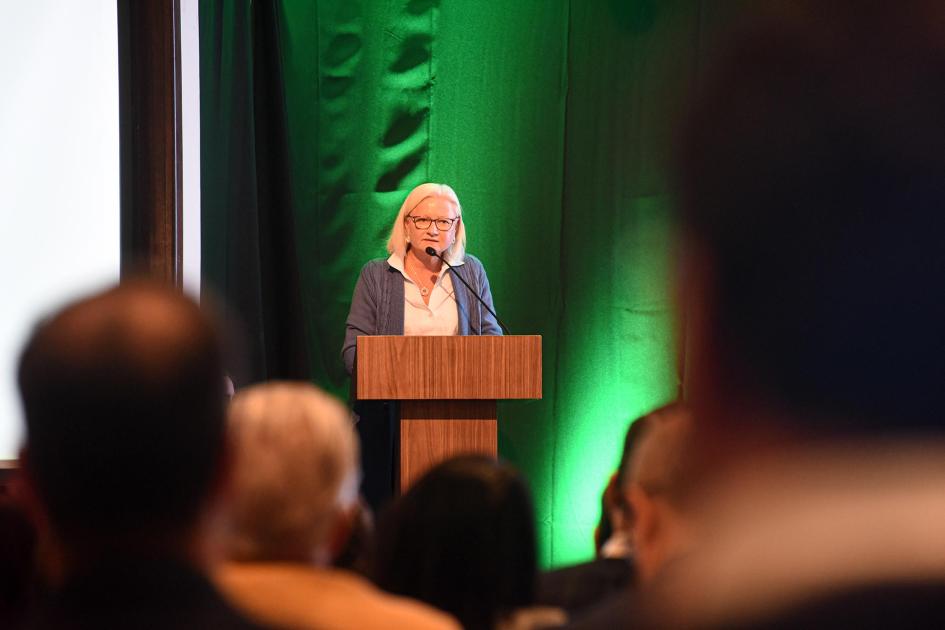 Representante del BID, Natacha Marzolf Representante del BID, Natacha Marzolf, habla desde un atril; en primer plano, público de espaldas