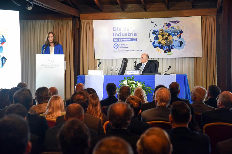 La ministra Elisa Facio fue una de las oradoras del evento Ministra Facio habla desde un atril; a su lado, en una mesa, está sentado Pache; al frente, púlblico