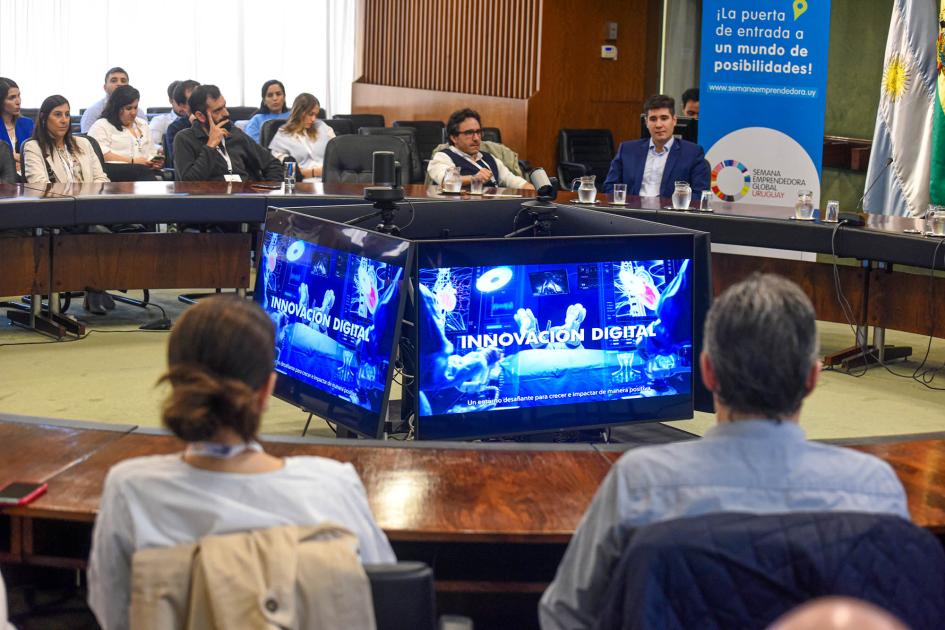 Público sentado en una mesa circular; en el medio se ven pantallas que forman un prisma