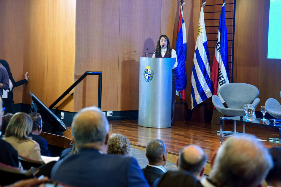 Disertación de la ministra Fernanda Cardona Cardona habla desde un atril; detrás, banderas; más cerca, público, de espaldas
