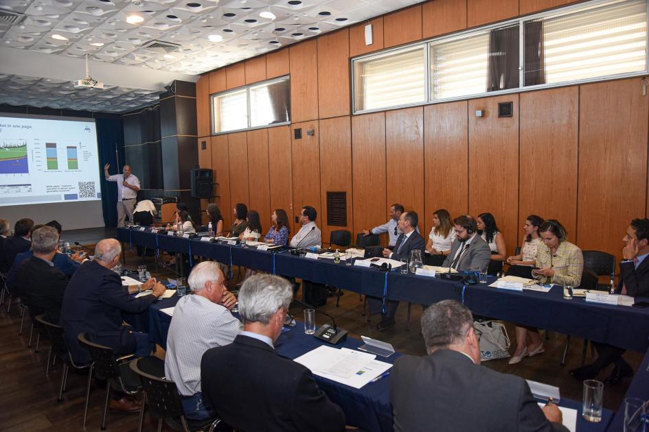 Grupo de personas, sentadas en mesas en forma de "u"; al fondo alguien habla desde una pantalla