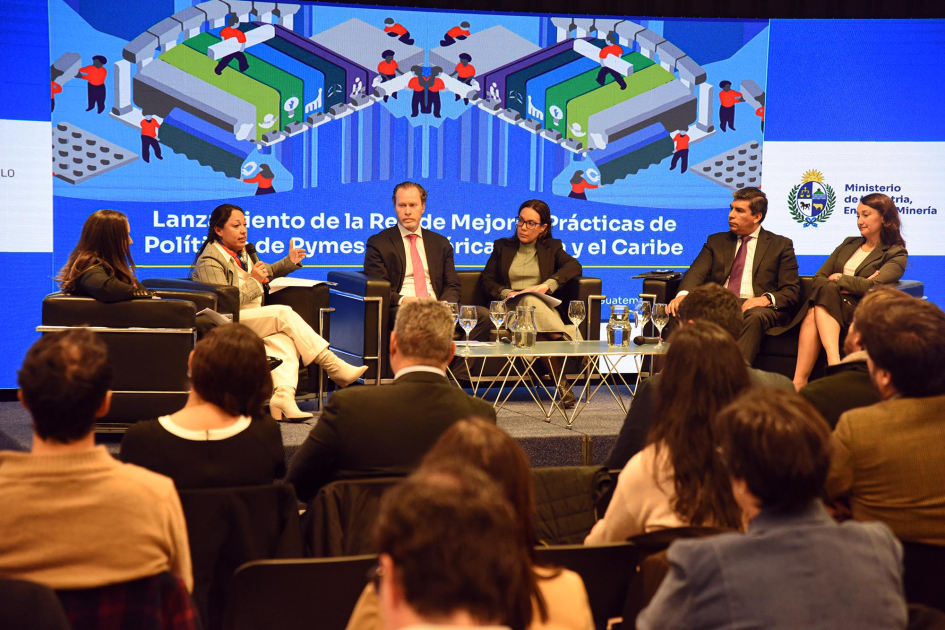 Al fondo, panelistas, sentados; más cerca, público, sentado y de espaldas