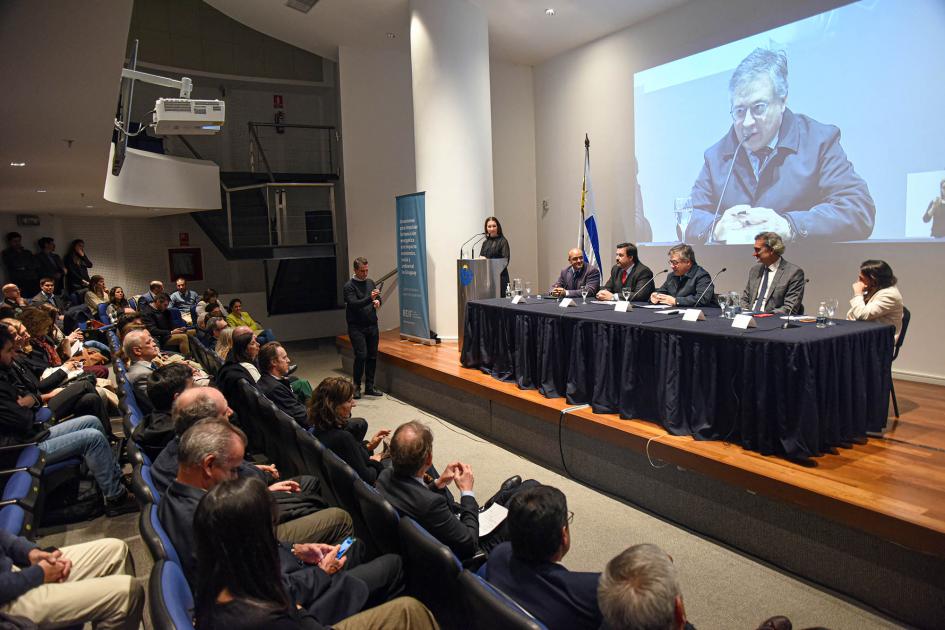 Mesa de autoridades y público presente Mesa de autoridades; habla Paganini y se lo ve en pantalla gigante, detrás; adelante, el público