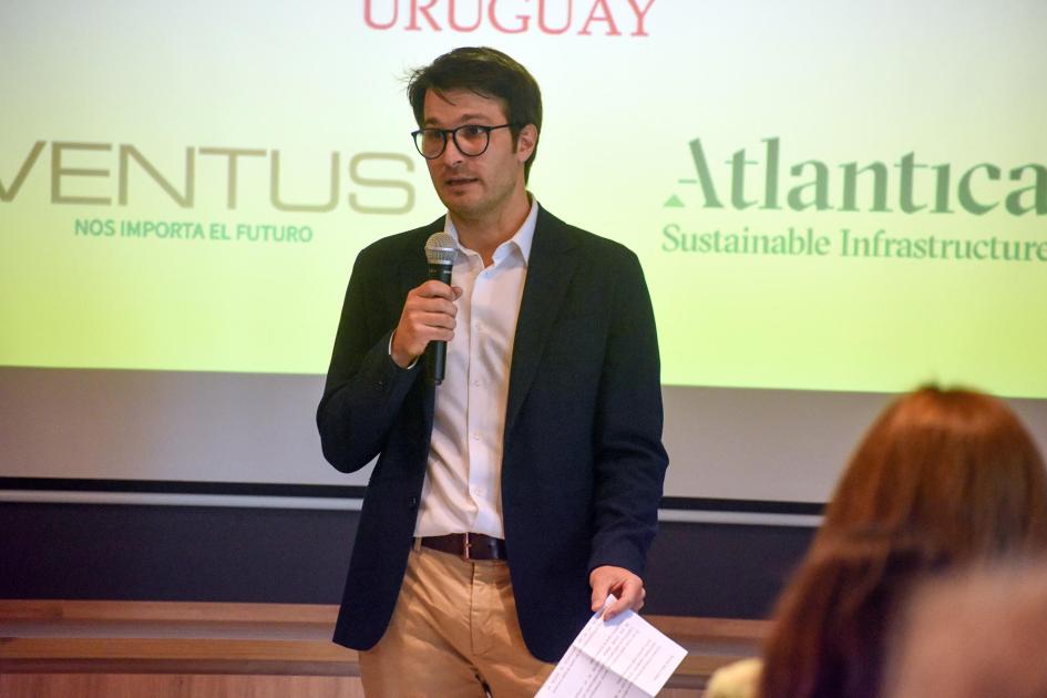 Un hombre habla con un micrófono; frente a él, de espaldas, el público; detrás, una pantalla