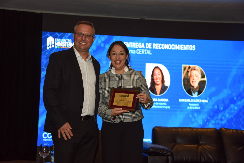 La ministra Fernanda Cardona recibió un reconocimiento de CERTAL Cardona, de pie junto a un representante de CERTAL; detrás, pantalla