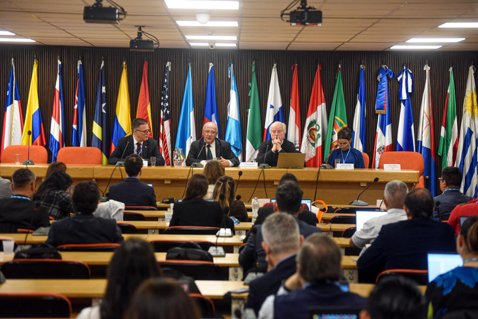 Capacitación sobre estadísticas subnacionales para medir la conectividad universal y significativa Al fondo, mesa de autoridades y banderas; más cerca, participantes, sentados de espaldas, en mesas