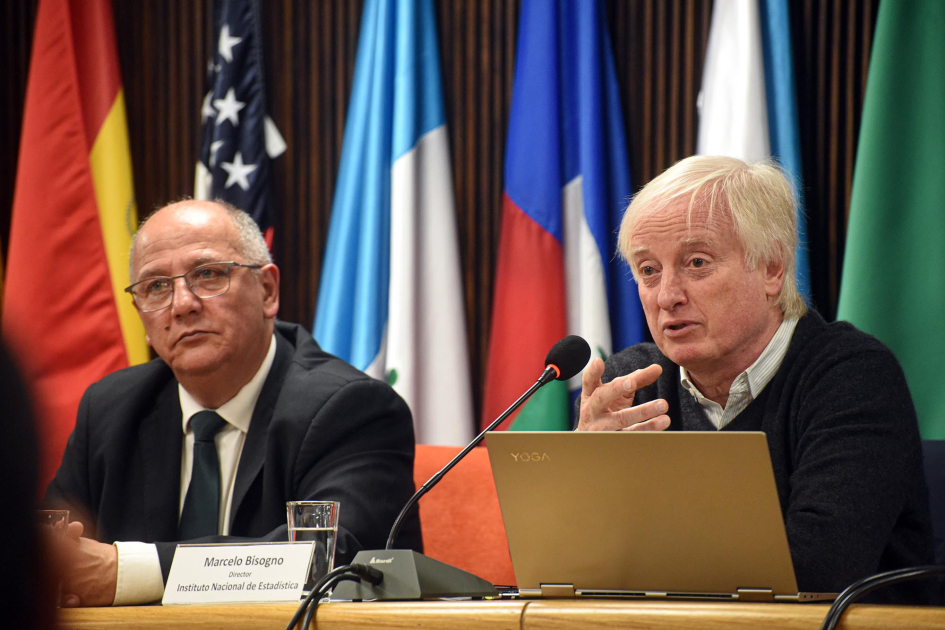 Director técnico del INE, Marcelo Bisogno Bisogno habla en una mesa, junto a Balseiro y con una laptop enfrente; detrás, banderas