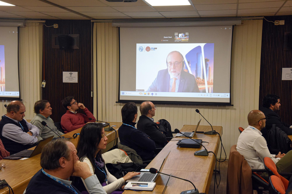 Bruno Ramos, director de la oficina regional de la UIT, participó a distancia Participantes, sentados en mesas; en la pared, imagen de Ramos