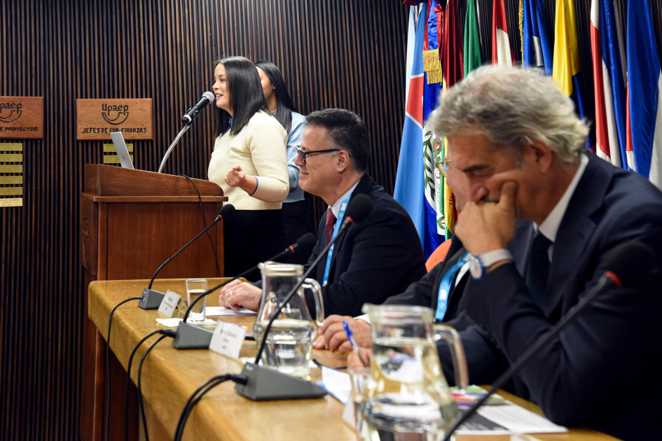 Blenda Silva, punto focal de la Juventud de UIT Américas, ofreció el saludo inicial en el evento En un atril, habla Silva, junto a la maestra de ceremonias; más cerca, autoridades en mesa