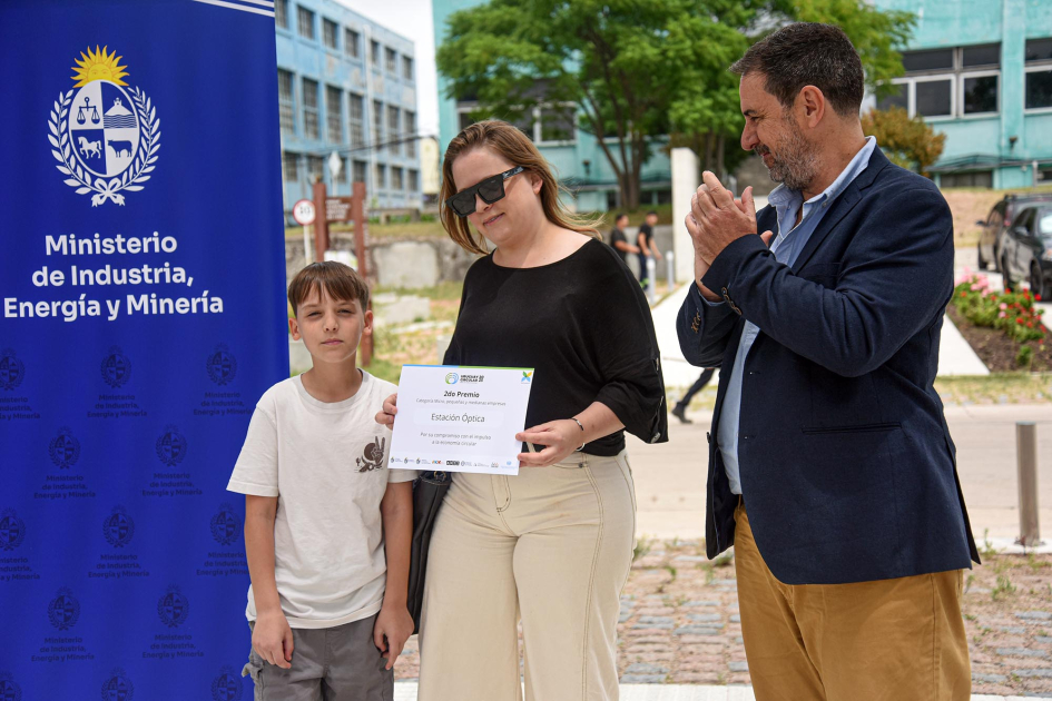 Una mujer sostiene un diploma junto a un niño y un banner del MIEM; al lado, aplaude un hombre