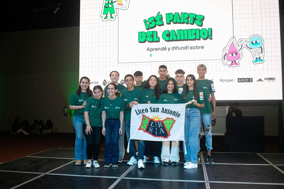 Adolescentes y docentes con una bandera que dice "Liceo Rural San Antonio"; detrás, una pantalla