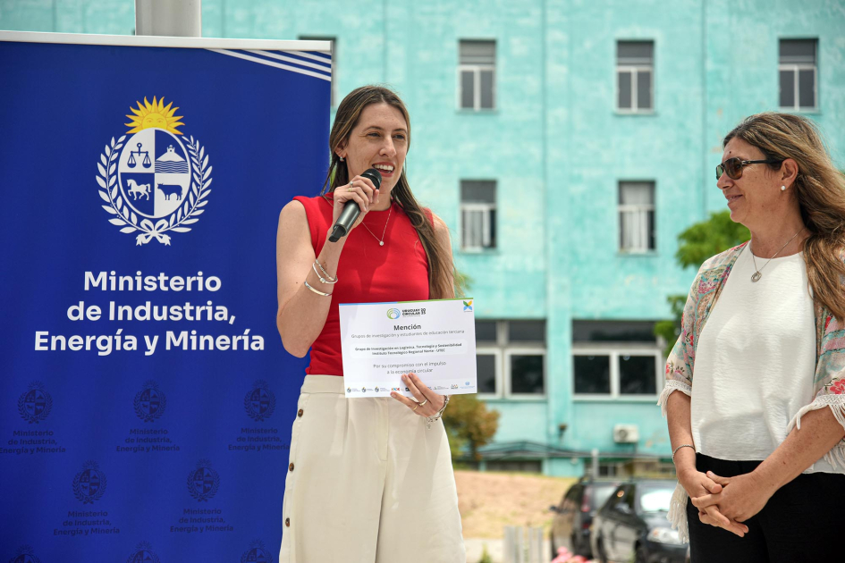 Mujer sostiene un diploma junto a otra mujer, mientras habla con micrófono