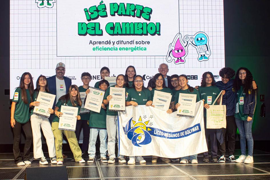 Liceo Médanos de Solymar Adolescentes y docentes con una bandera que dice "Liceo Médanos de Solymas"; detrás hay una pantalla