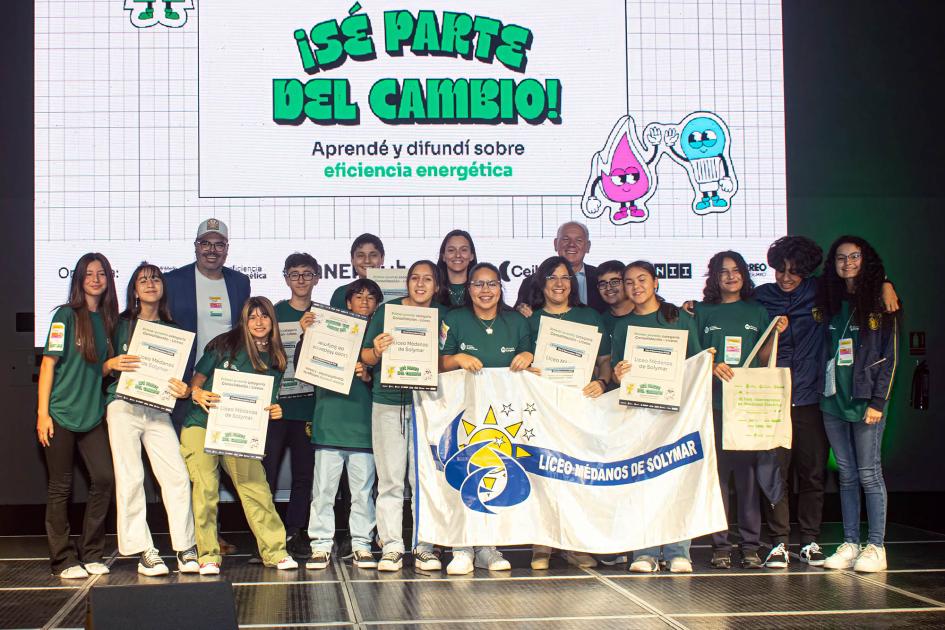 Adolescentes y docentes con una bandera que dice "Liceo Médanos de Solymas"; detrás hay una pantalla