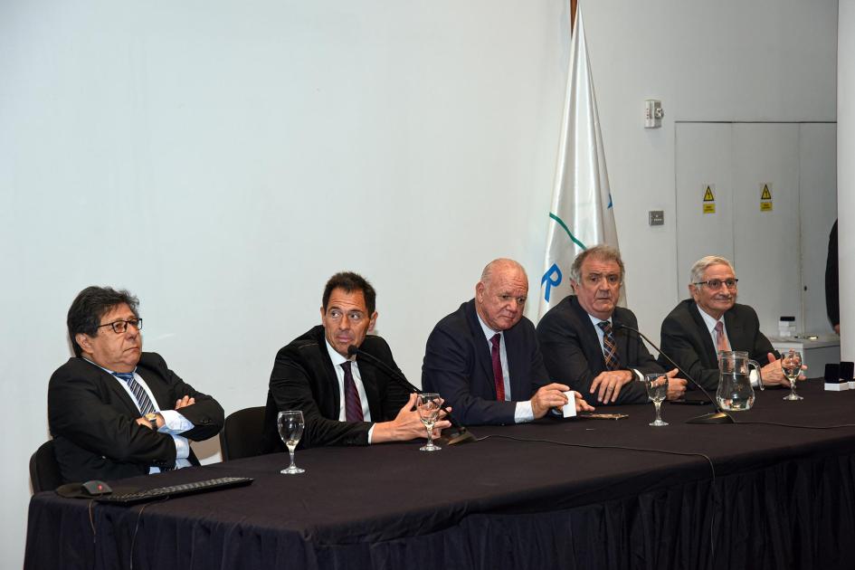 Mesa de autoridades (cinco hombres); al lado, bandera del Mercosur