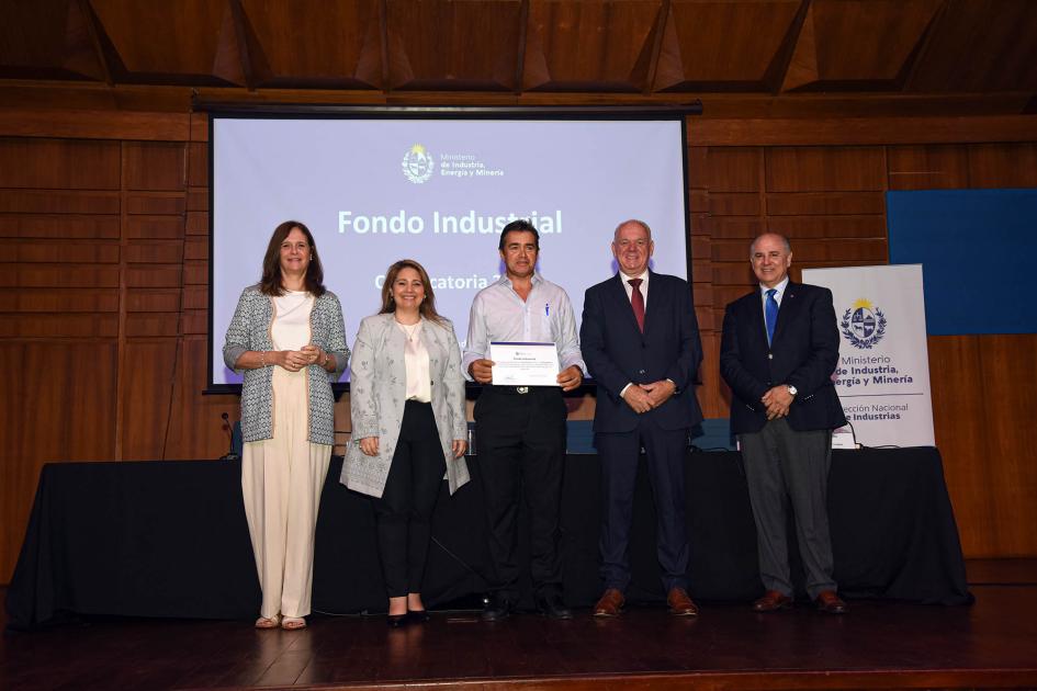 Autoridades acompañan a un hombre que muestra su diploma; detrás hay una pantalla